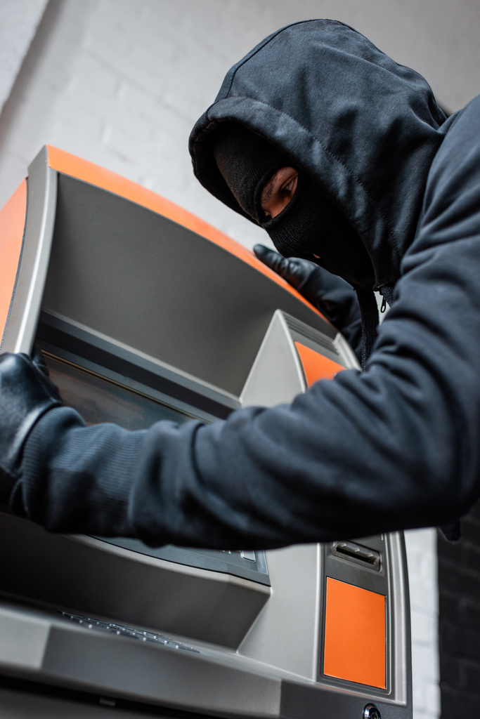 Tiefer Blickwinkel auf Einbrecher mit Sturmhaube und Schaum-Handschuhen, die Geldautomaten halten - Foto, Bild