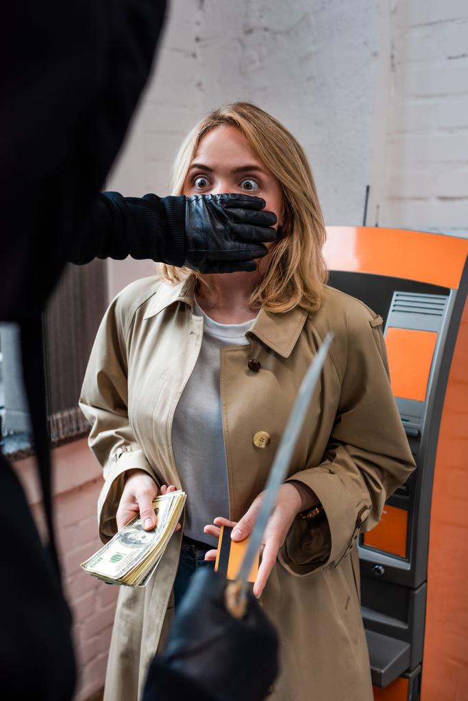 Selektivní zaměření lupiče s nožem zakrývající ústa na vyděšenou ženu držící kreditní kartu a peníze  - Fotografie, Obrázek