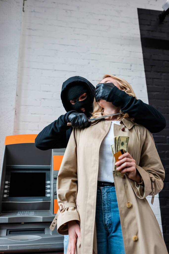 Rabló kést tart a lány nyaka közelében, kezében dollár és hitelkártya mellett ATM  - Fotó, kép