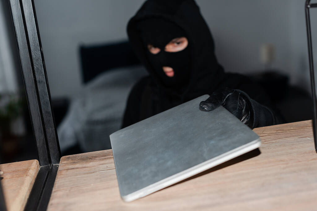 Foco seletivo de ladrão em balaclava e luva de couro levando laptop do armário
  - Foto, Imagem