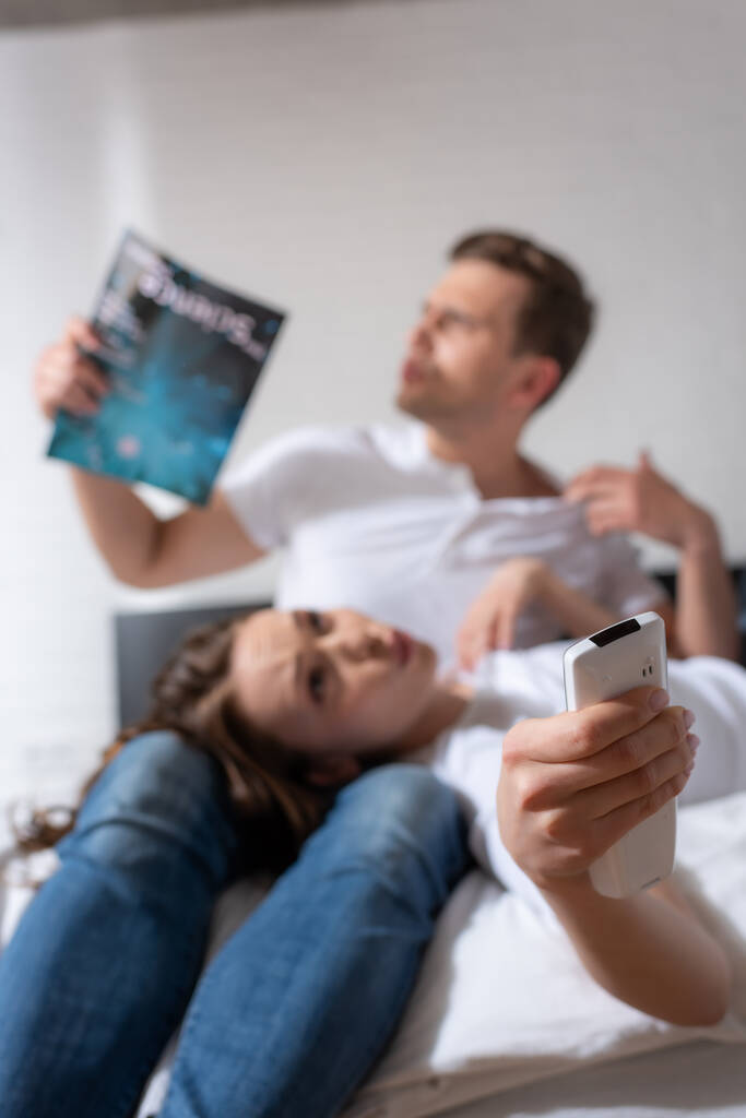 selektiver Fokus des Mädchens mit Fernbedienung von einer Klimaanlage, die in der Nähe eines Mannes liegt, der ein Magazin hält, während er sich heiß fühlt  - Foto, Bild