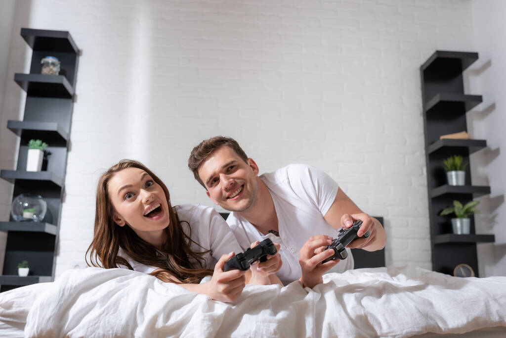 KYIV, UCRANIA - 15 de mayo de 2020: mujer alegre y hombre feliz acostado en la cama y jugando videojuegos
  - Foto, imagen