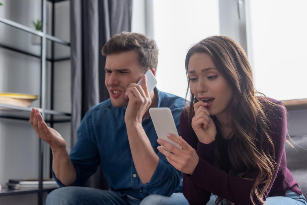selective focus of surprised girl looking at smartphone while displeased man talking on smartphone in living room  - Photo, Image