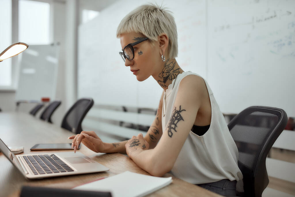 Moderni bisnesnynny. Sivunäkymä nuori ja kaunis tatuoitu nainen, jolla on lyhyt kampaus kannettavan tietokoneen avulla, kun hän istuu työpaikallaan kokoushuoneessa - Valokuva, kuva