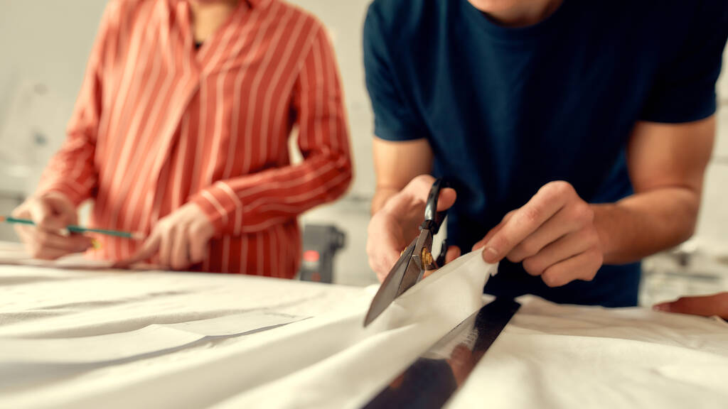 Iets unieks creëren. Gesneden foto van jonge mannelijke ontwerper snijden witte stof textiel in een studio. Groep van creatieve millennials die samenwerken - Foto, afbeelding