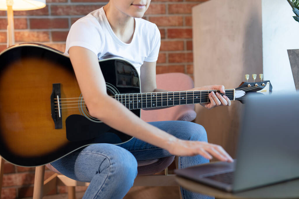 Jeune femme assise dans une pièce sur un fauteuil, apprend à jouer de la guitare en ligne. Formation en ligne, cours de musique en ligne, formation vocale et guitare en ligne. Reste à la maison
. - Photo, image