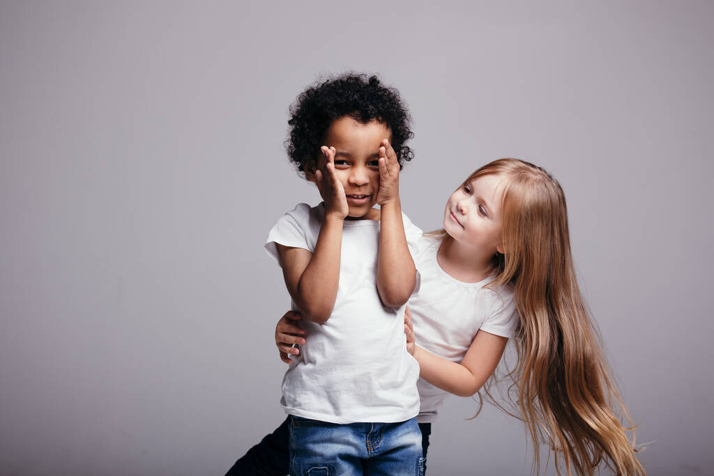Pequena menina de cabelos compridos fica atrás de um menino afro-americano encaracolado que cobre o rosto com as mãos em um fundo cinza
 - Foto, Imagem