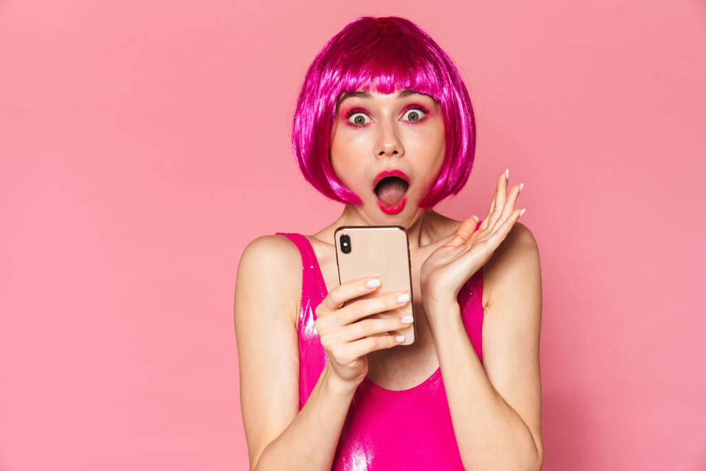 Imagen de la chica de la fiesta con peluca expresando sorpresa y usando teléfono móvil aislado sobre fondo rosa
 - Foto, Imagen
