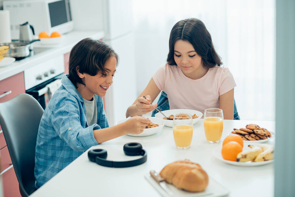Pozitivní chlapec a dívka sedí u kuchyňského stolu s miskami kukuřičných vloček a sklenicemi pomerančového džusu - Fotografie, Obrázek