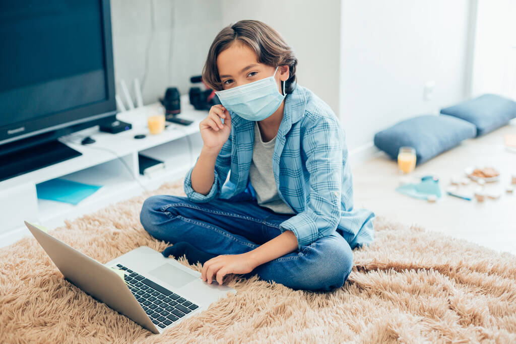 Niño tranquilo en ropa casual con una máscara médica mientras está sentado en la cama delante de una pantalla de ordenador portátil
 - Foto, imagen