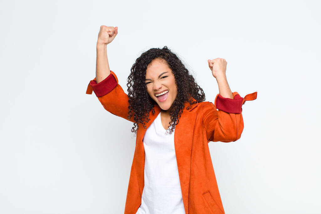 νεαρή μαύρη γυναίκα να φωνάζει θριαμβευτικά, δείχνοντας ενθουσιασμένη, χαρούμενη και έκπληκτη νικήτρια, γιορτάζοντας ενάντια στον λευκό τοίχο - Φωτογραφία, εικόνα