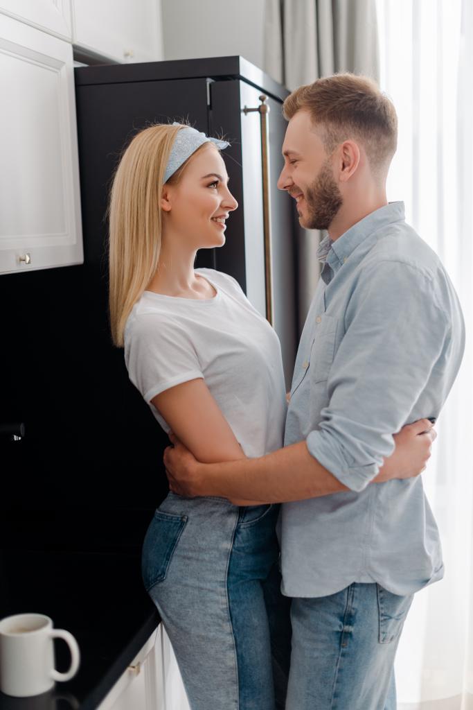 Mutlu kız arkadaş ve sakallı erkek arkadaş mutfakta sarılırken birbirlerine bakıyorlar.  - Fotoğraf, Görsel