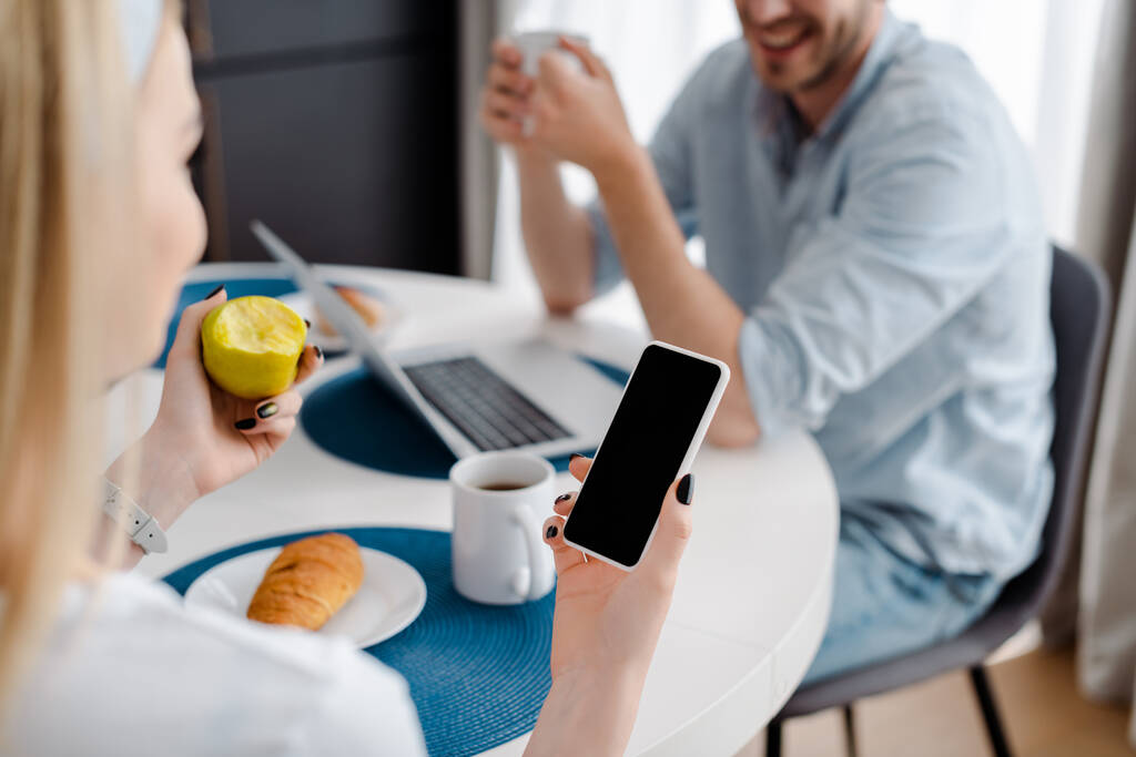 foco seletivo da mulher segurando smartphone com tela em branco e maçã perto freelancer alegre e café da manhã
  - Foto, Imagem