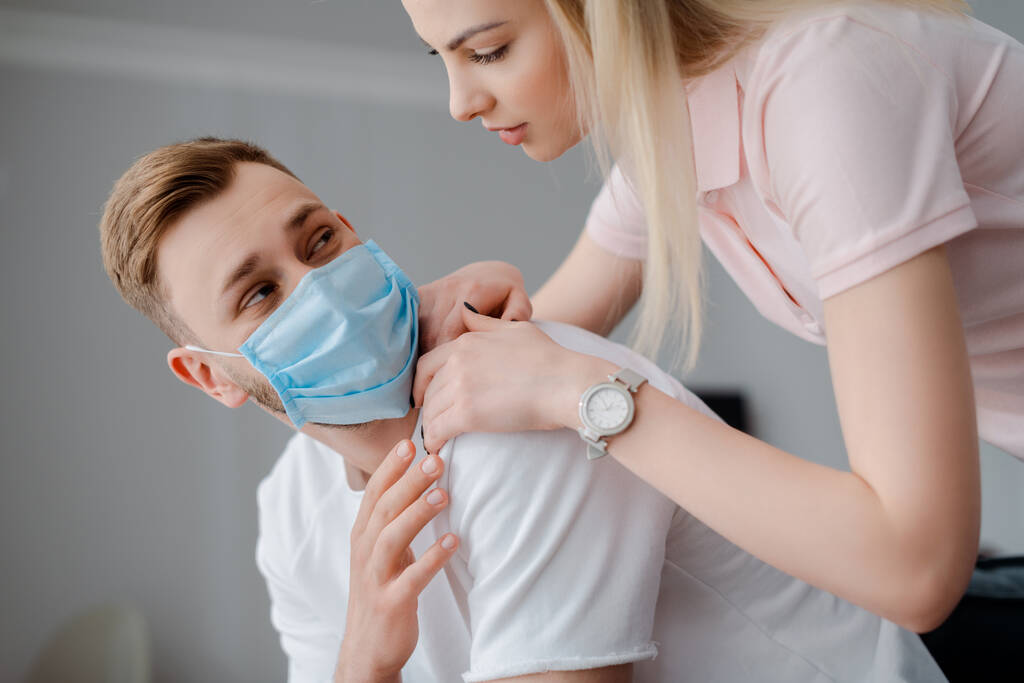 hermosa chica tocando frustrado hombre en máscara médica en casa
  - Foto, Imagen