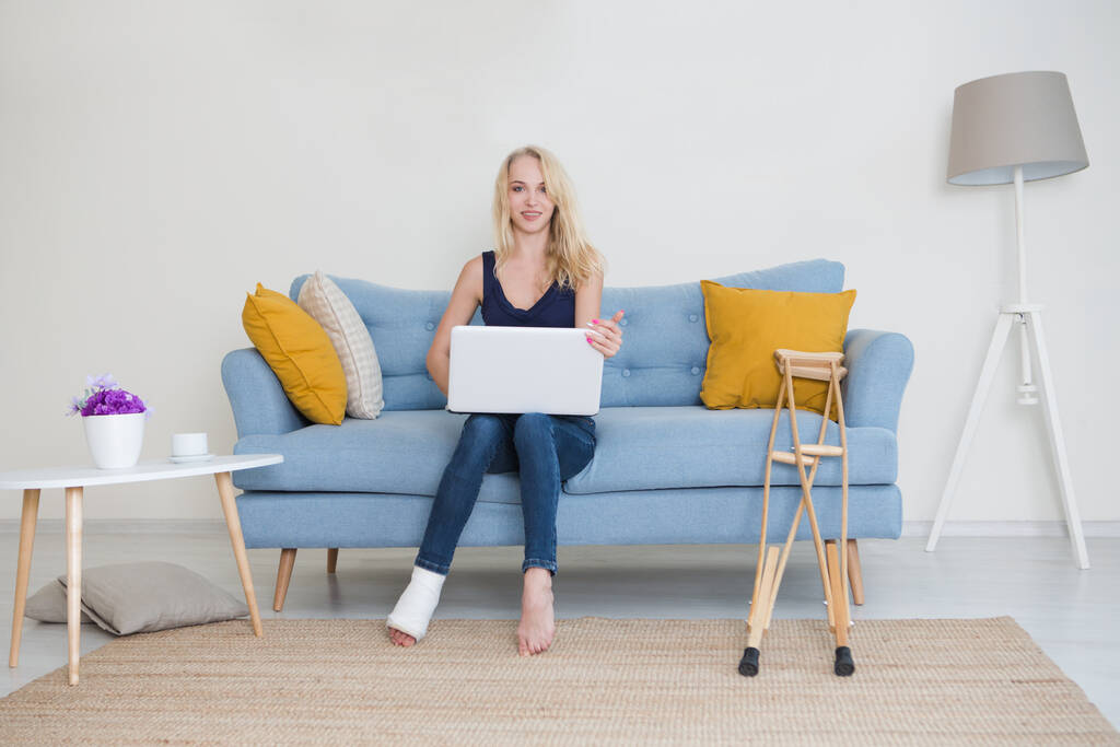 Młoda kobieta ze złamaną nogą w gipsie, siedząca na kanapie w domu i pracująca na laptopie. Chora kobieta rozmawia z lekarzem online. konsultacje z lekarzem online - Zdjęcie, obraz