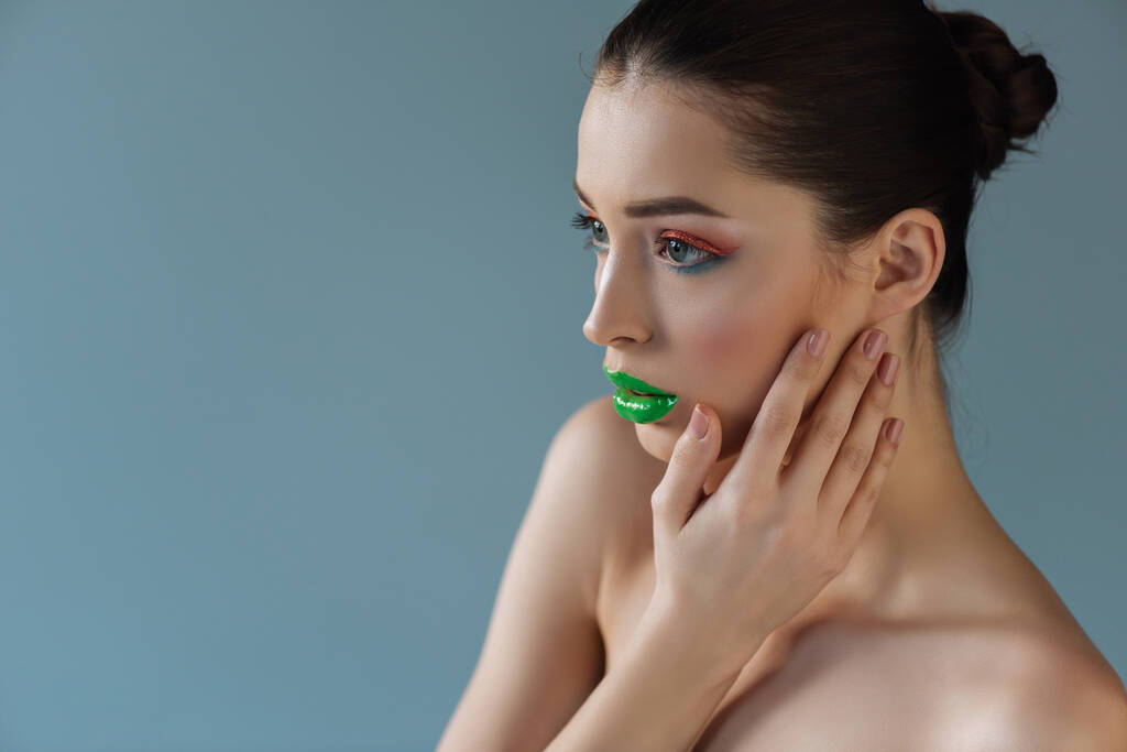 nue belle femme aux lèvres vertes, fard à paupières rose et bleu touchant visage isolé sur bleu - Photo, image