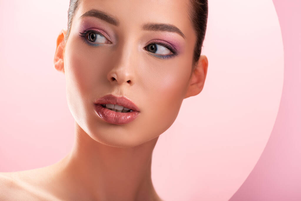 портрет красивой обнаженной женщины с блестящим макияжем, смотрящей сквозь круглые бумажные отверстия, изолированные на розовый
 - Фото, изображение