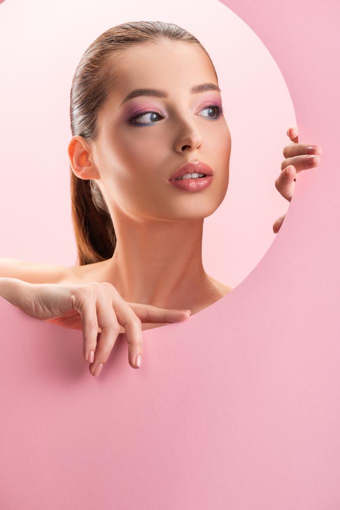 portrait of beautiful naked woman with shiny makeup looking through round paper hole isolated on pink - Photo, Image