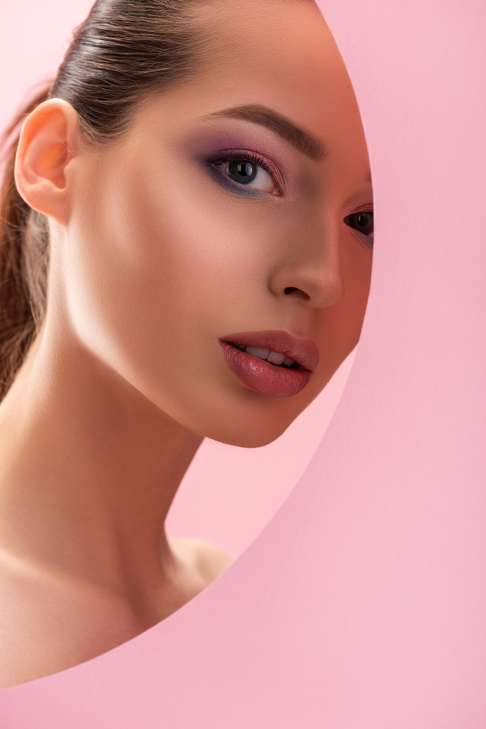 Porträt einer schönen nackten Frau mit glänzendem Make-up, die durch ein rundes Papierloch auf rosa isoliert blickt - Foto, Bild