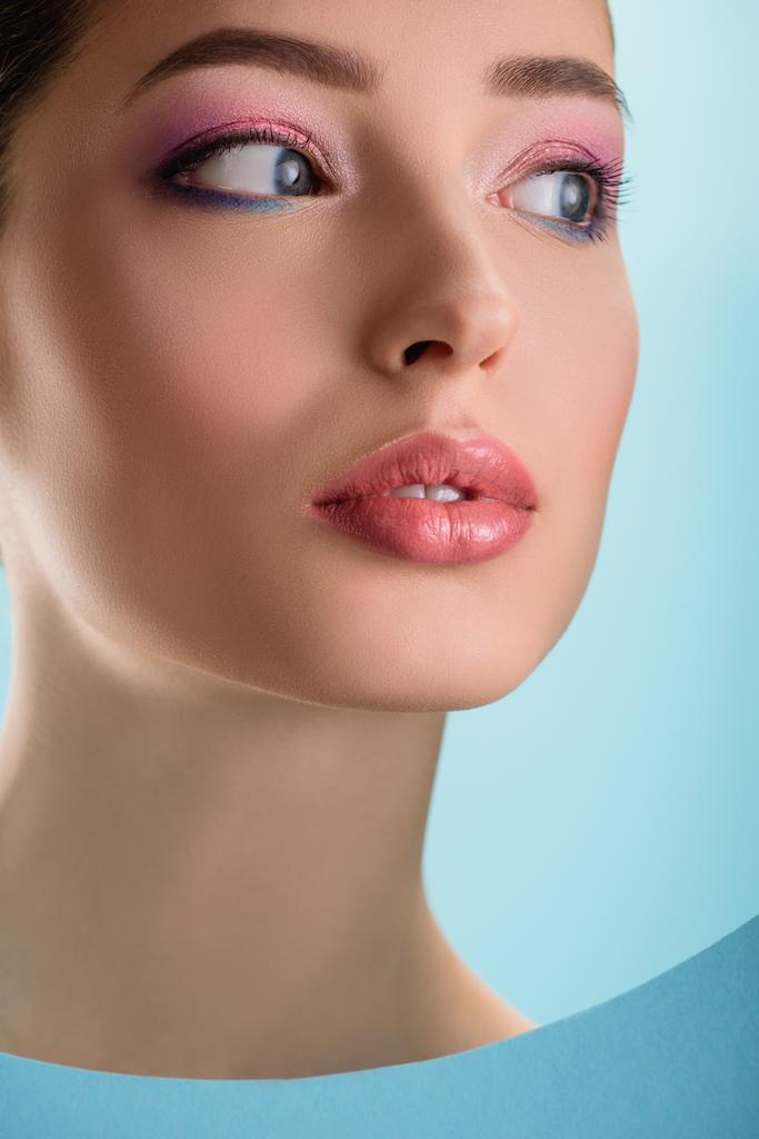 πορτρέτο της όμορφης γυναίκας με γυαλιστερά χείλη, ροζ σκιά ματιών κοιτάζοντας μέσα από χαρτί στρογγυλή τρύπα απομονωμένη στο μπλε - Φωτογραφία, εικόνα