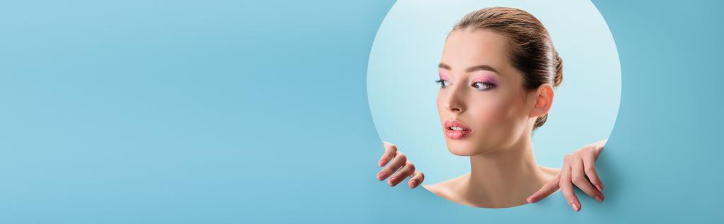 portrait of beautiful naked woman with glossy lips, pink eyeshadow looking away through paper round hole isolated on blue, panoramic shot - Photo, Image