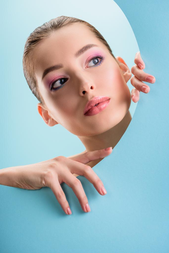 retrato de mulher nua bonita com lábios brilhantes, sombra de olhos rosa olhando através de papel buraco redondo isolado em azul
 - Foto, Imagem