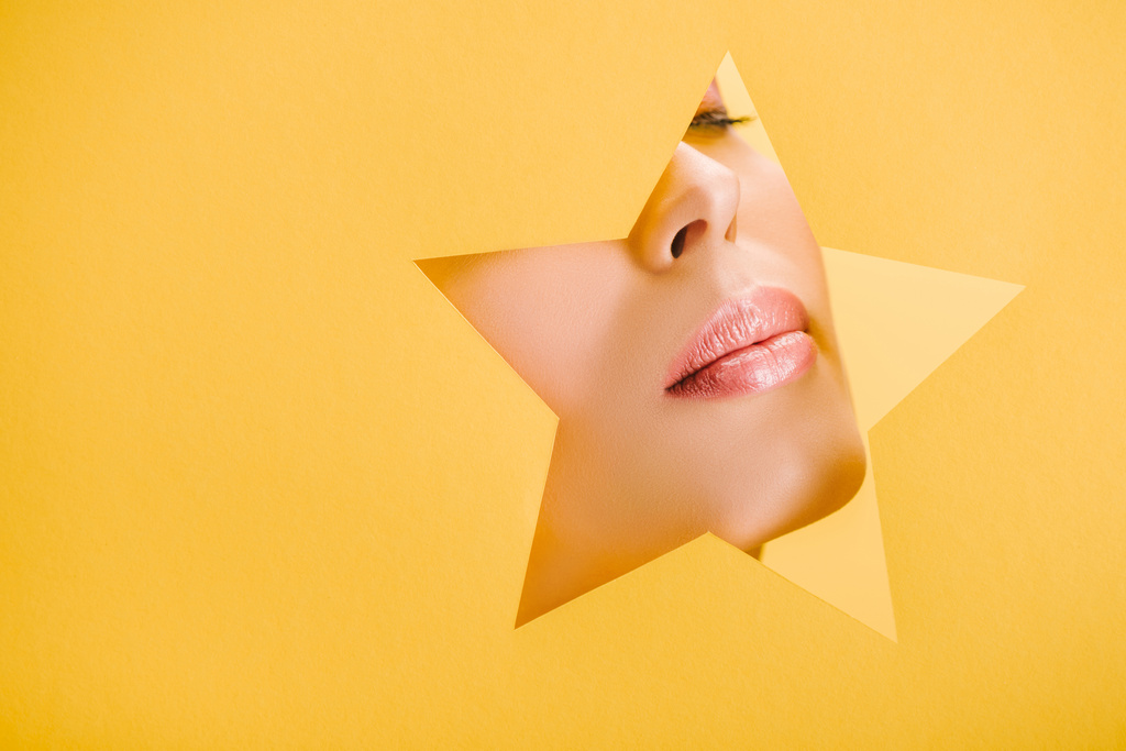 abgeschnittene Ansicht der schönen Frau mit glänzenden Lippen in Papier sternförmigen Loch isoliert auf gelb - Foto, Bild