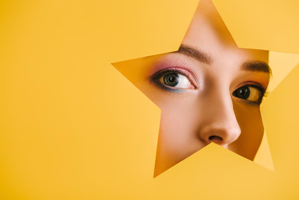 portret pięknej kobiety z błyszczącym makijażem w papierowej dziurze w kształcie gwiazdy patrząc na aparat odizolowany na żółto - Zdjęcie, obraz