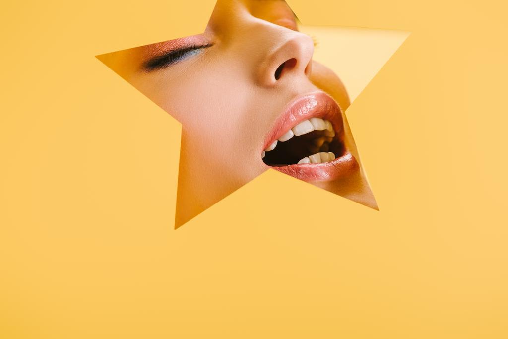 portrait of beautiful woman with shiny makeup in paper star shaped hole with closed eyes and open mouth isolated on yellow - Photo, Image