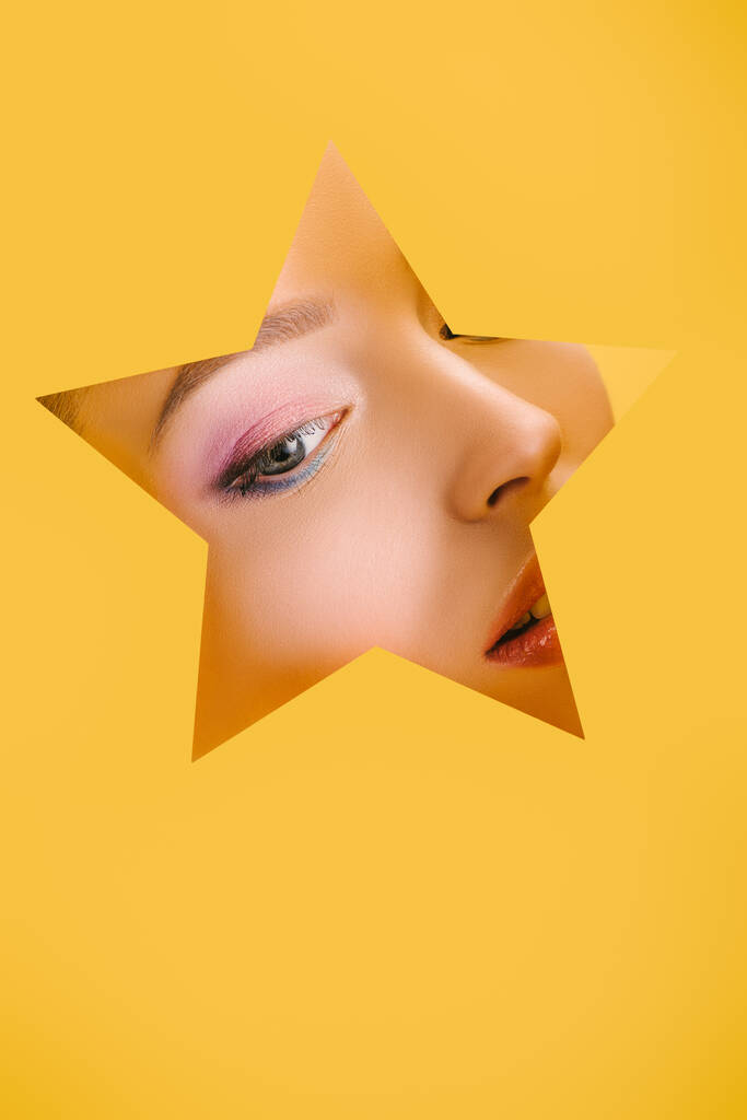 Kağıt yıldız şeklindeki delikte parlak makyajlı güzel bir kadının portresi sarıda izole bir şekilde duruyor. - Fotoğraf, Görsel