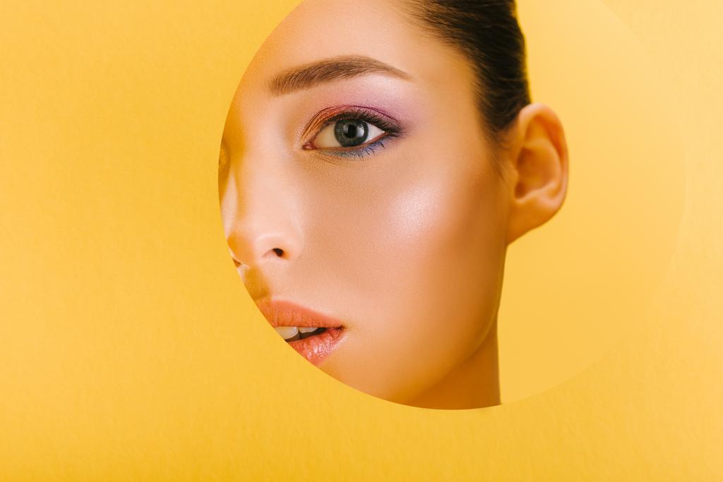 portrait of beautiful woman with shiny makeup in paper round hole looking at camera isolated on yellow - Photo, Image