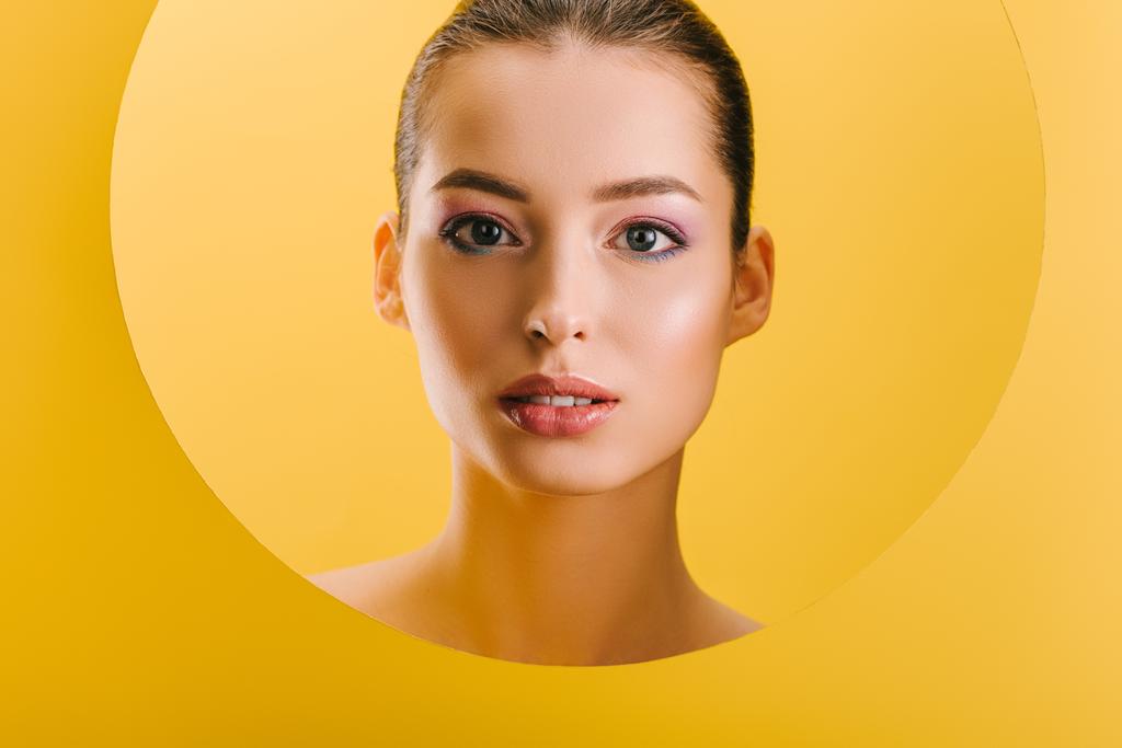 portrait of beautiful woman with shiny makeup in paper round hole looking at camera isolated on yellow - Photo, Image