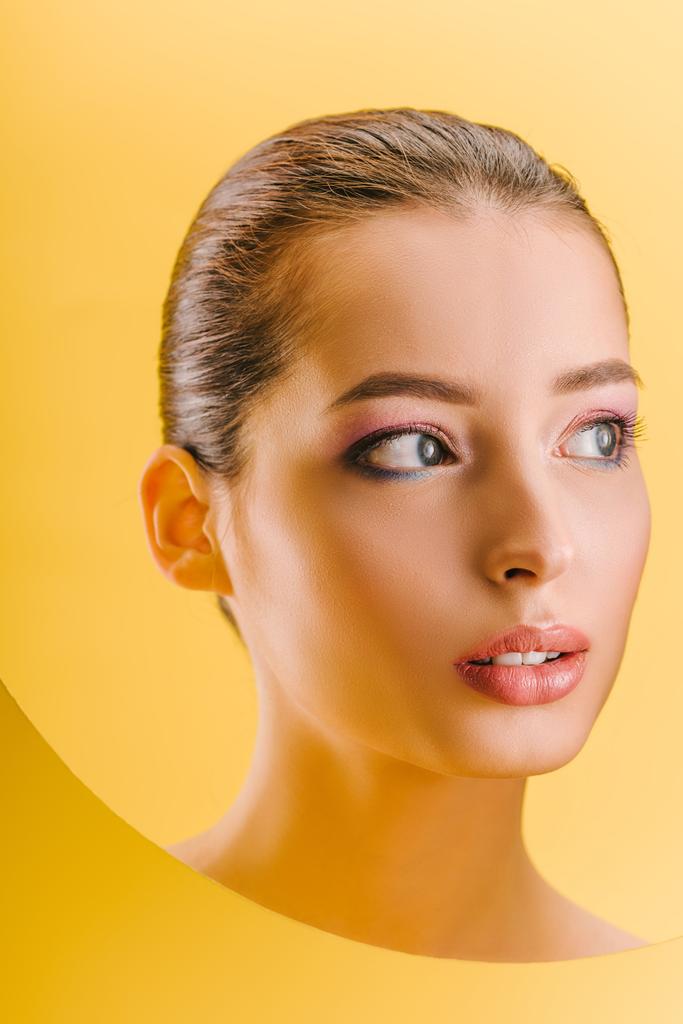 Porträt einer schönen Frau mit glänzendem Make-up in einem runden Loch aus Papier, das isoliert auf gelb wegschaut - Foto, Bild