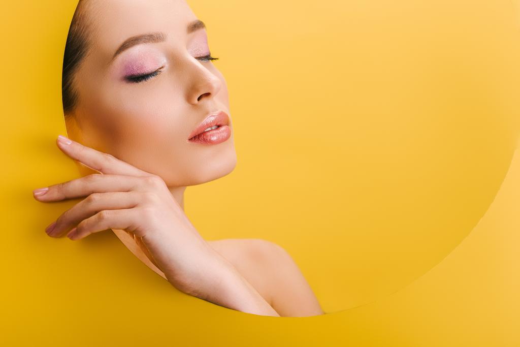 Porträt der schönen nackten Frau mit glänzendem Make-up in Papier runde Loch mit der Hand und geschlossenen Augen isoliert auf gelb - Foto, Bild