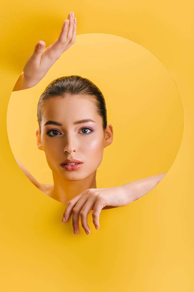 portrait of beautiful woman with shiny makeup in paper round hole with hands isolated on yellow - Photo, Image