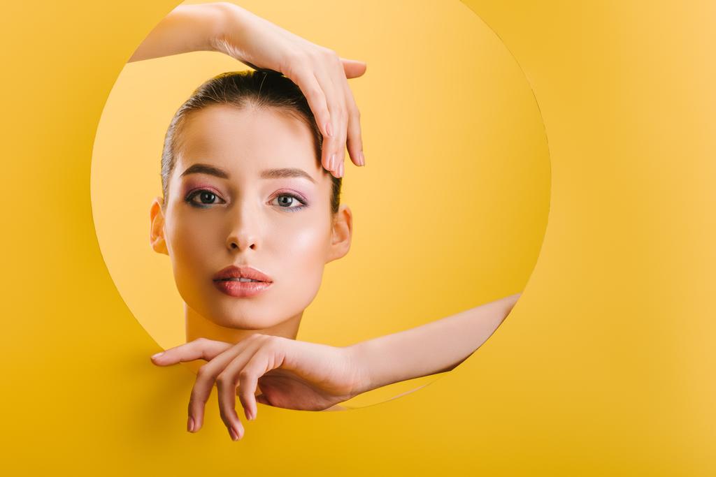 Porträt der schönen Frau mit glänzendem Make-up in Papier runde Loch mit den Händen isoliert auf gelb - Foto, Bild