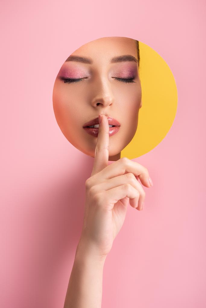 portrét krásné ženy s lesklým make-upem a zavřenýma očima v růžovém papíru kulatý otvor ukazující shh znamení izolované na žluté - Fotografie, Obrázek