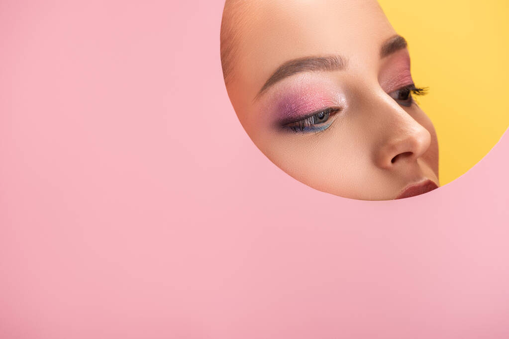 portrait of beautiful woman with shiny eyeshadow in pink paper round hole looking away isolated on yellow - Photo, Image