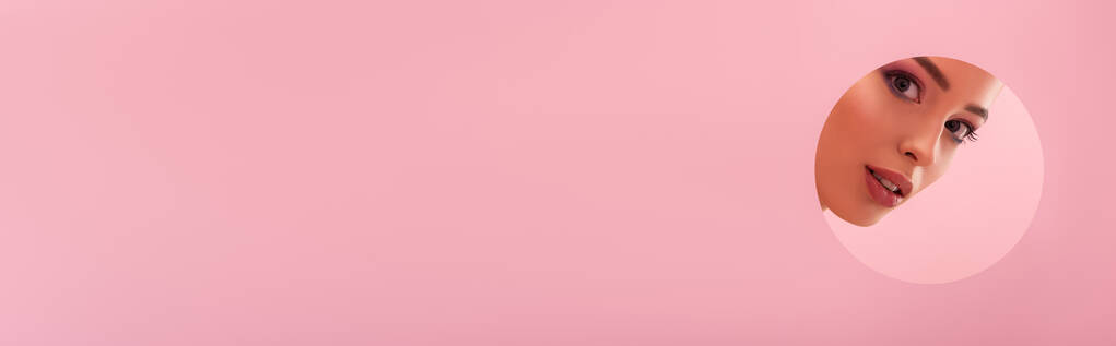 retrato de mulher bonita com maquiagem brilhante em papel buraco redondo isolado em rosa, tiro panorâmico
 - Foto, Imagem