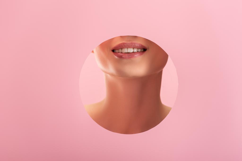 Teilansicht der schönen lächelnden Frau mit glänzenden Lippen und weißen Zähnen in einem auf rosa Papier isolierten runden Loch - Foto, Bild