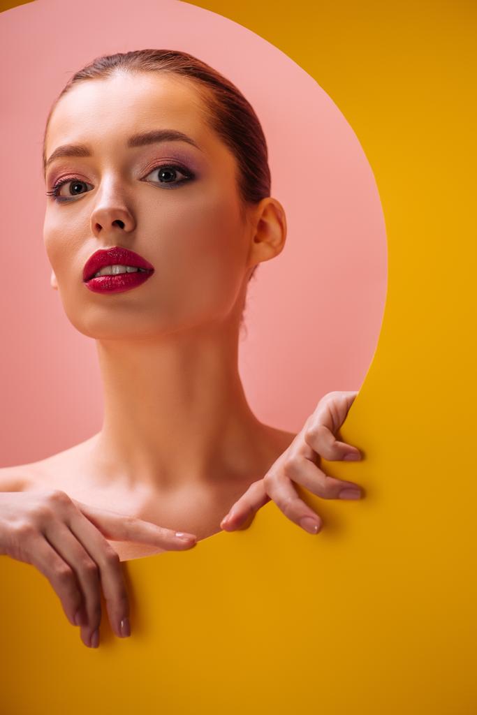 nackte schöne Frau mit purpurroten Lippen in gelbem Papierkreisloch auf rosa Hintergrund - Foto, Bild