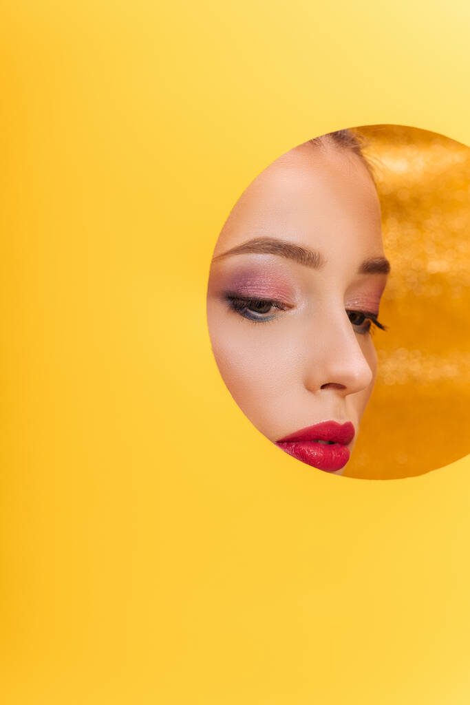 belle femme avec un maquillage coloré regardant à travers le trou de cercle en papier jaune - Photo, image