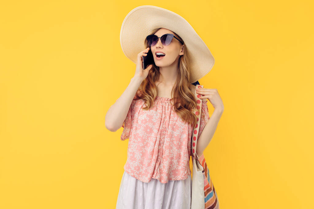 Joyeux sourire jeune femme dans un chapeau d'été et des lunettes de soleil, parler sur un téléphone portable sur un fond jaune isolé
 - Photo, image