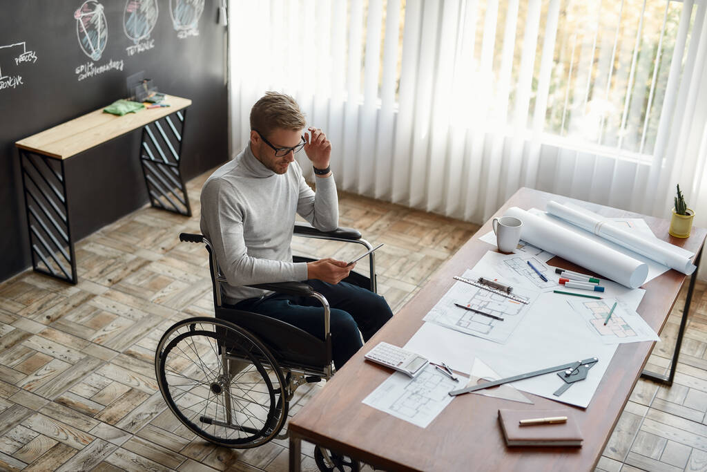 Luogo di lavoro. Ingegnere o architetto maschio concentrato su una sedia a rotelle che lavora con tablet digitale sul posto di lavoro nel suo ufficio moderno
 - Foto, immagini