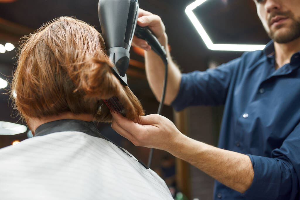 Kapsel aan het maken. Jonge professionele kapper drogen lange roodharige haar van zijn klant in kapperszaak - Foto, afbeelding