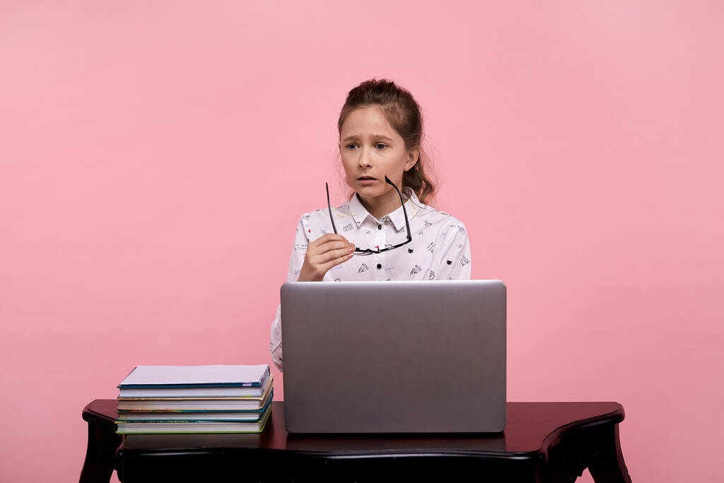 Una ragazza sconvolta siede a un computer portatile con una camicia bianca e tiene gli occhiali con cornice nera. - Foto, immagini
