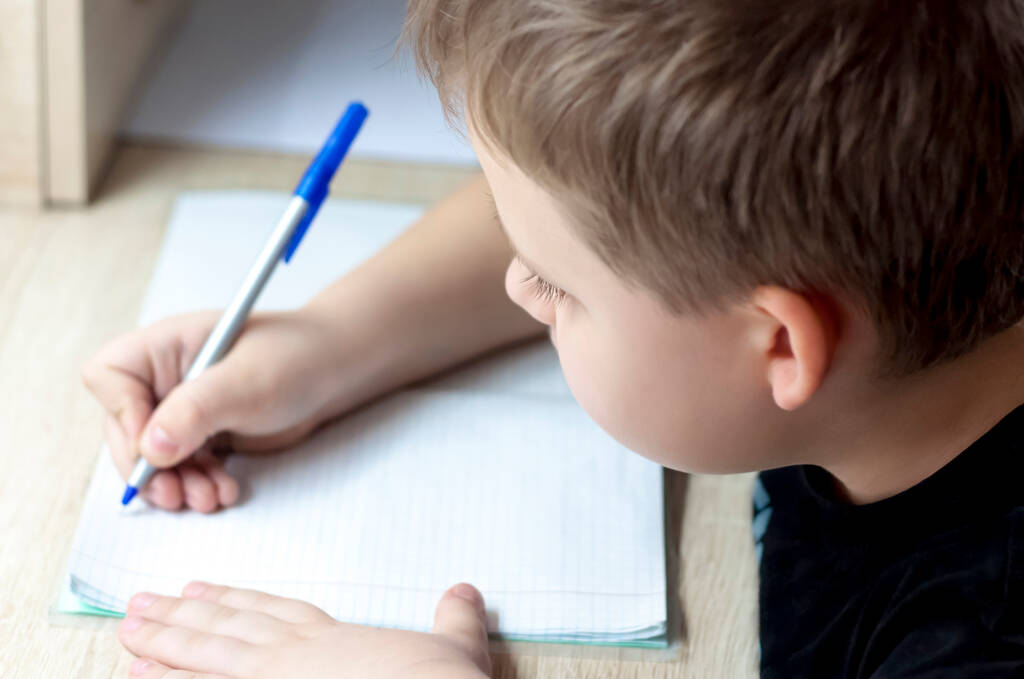 Εκπαίδευση στο σπίτι. Το αγόρι κάνει τα μαθήματά του. Γράφει σε ένα σημειωματάριο. - Φωτογραφία, εικόνα