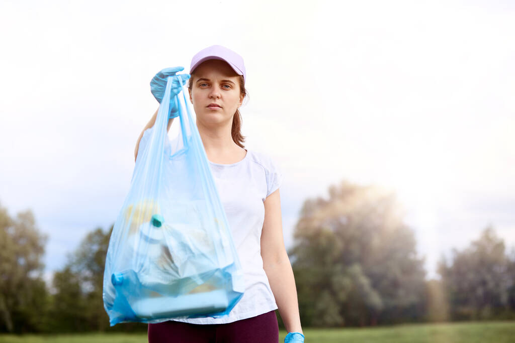 Nuori valkoihoinen tyttö yllään t paita ja baseball-lippis poseeraa ulkona roskapussi, välittää ympäristöstä, seisoo taivaalla ja puita taustalla. - Valokuva, kuva