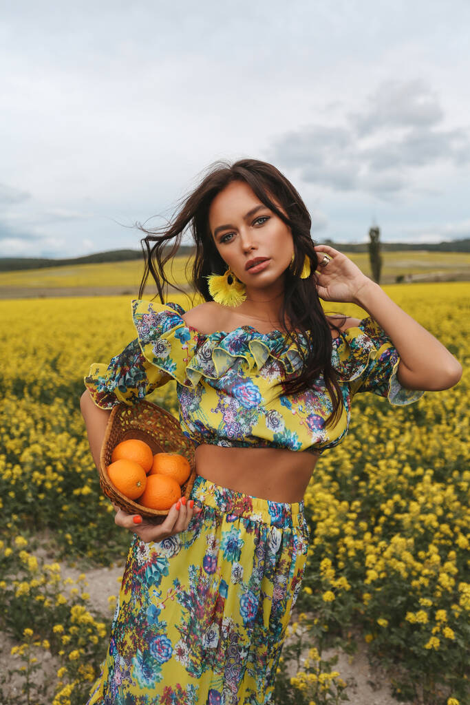 photo de mode de belle femme sexy aux cheveux foncés dans des vêtements élégants posant dans le champ avec des fleurs à fleurs jaunes, tenant des oranges dans le panier - Photo, image
