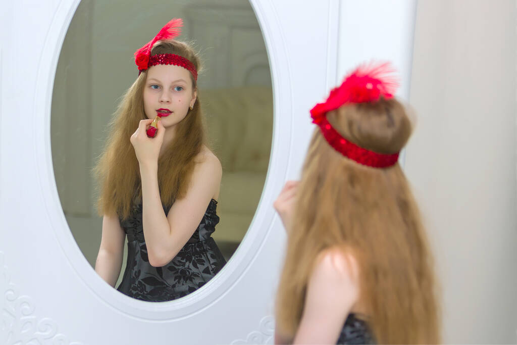 Piękna dziewczyna w wieku szkolnym maluje usta szminką i patrzy w lustro. - Zdjęcie, obraz